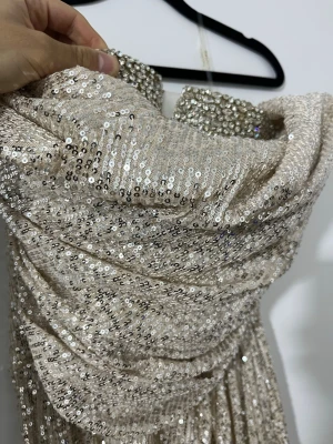 Champagnefärgad paljettklänning - Snygg axelbandslös långklänning i champagnefärg med glittriga paljetter över hela tyget. Klänningen har vadderad byst och en elegant draperad design. Perfekt för dig som vill sticka ut på bal eller fest.