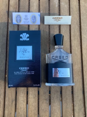 Creed Aventus Eau de Parfum 100ml - Creed Aventus Eau de Parfum 100ml med elegant glasflaska och svart etikett. Parfymen levereras i originalförpackning med tillhörande informationskort. Flaskan har en stilren design med silverdetaljer och svart kork.