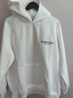 Vit hoodie Essentials Fear of God - Snygg vit hoodie från Essentials Fear of God med svart logga på bröstet. Klassisk känguruficka framtill och stor huva. Tillverkad i mjukt bomullsmaterial som känns riktigt skönt mot huden. Perfekt för en clean och stilren streetwear-look.