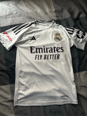 Real Madrid matchtröja Adidas vit - Säljer en vit Real Madrid matchtröja från Adidas med svarta detaljer och klubbmärke på bröstet. Tröjan har tryckta sponsorer, La Liga-logga på ärmen och klassiska Adidas-ränder på axlarna. Tillverkad i lätt och ventilerande material.