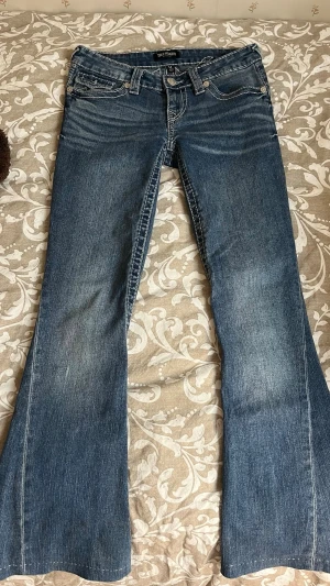 Blå bootcut jeans från True Religion - Säljer ett par blå utsvängda jeans från True Religion med snygga broderade detaljer och fickor bak. Jeansen har klassisk femficksmodell och coola kontrastsömmar. Förkortade för hand med handsydd uppläggning. Innerbenslängd ca 73 cm. 