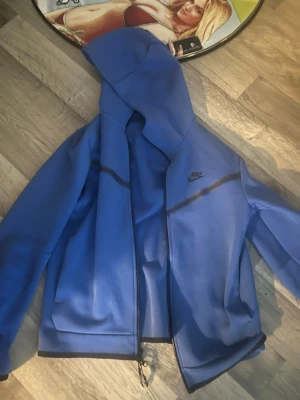 Blått Nike tech set - Säljer ett blått loungewear set från Nike med zip-hoodie och matchande byxor. Hoodien har dragkedja, fickor och svart Nike-logga på bröstet. Byxorna har dragsko i midjan och samma logga på benet. Perfekt för chill dagar hemma.