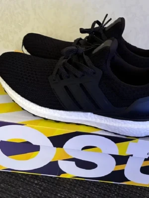 Adidas Ultra Boost svarta sneakers - Svarta Adidas Ultra Boost sneakers med stickad ovandel, vita Boost-mellansulor och svarta detaljer. Skorna har klassisk låg profil, snörning och en flexibel yttersula med grepp. Perfekta för dig som gillar sportig och stilren look.
