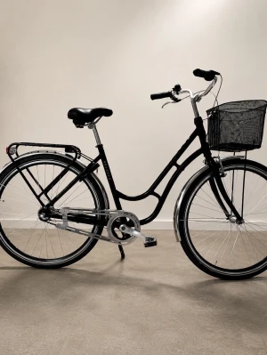 Svart damcykel med korg - Snygg svart damcykel med klassisk ram, svart sadel och praktisk korg framtill. Utrustad med pakethållare bak och reflexer på hjulen. Perfekt för att ta sig runt i stan eller till skolan.