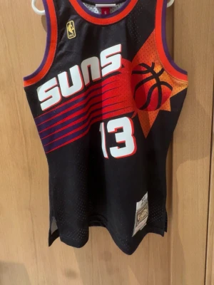 Phoenix Suns basketlinne Nash #13 - Snyggt svart basketlinne från Mitchell & Ness med Phoenix Suns-logga och Nash #13. Linnet har lila och orange detaljer, stora tryck fram och bak samt meshmaterial som andas. Perfekt för basketträning eller att bära som streetwear.