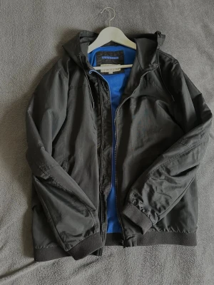 Svart vindjacka från Calvin Klein Jeans - Stilren svart vindjacka från Calvin Klein Jeans med huva och dragkedja framtill. Jackan har ribbade muddar vid ärmslut och nederkant samt meshfoder i blått för extra komfort. Perfekt för blåsiga dagar och enkel att matcha med olika outfits.