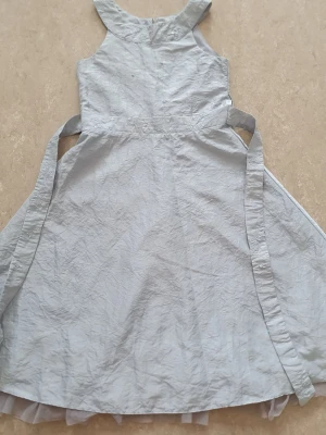 Klänning - Söt ljusgrå klänning med broderade detaljer och små paljetter i midjan. Klänningen har knytband i sidan och volangkant nertill. Perfekt för sommarens alla tillfällen. Fläck och hällorna är trasiga.