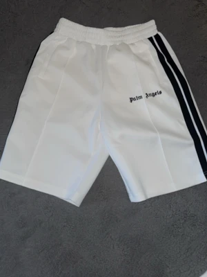 Vita Palm Angels shorts med svarta ränder - Säljer ett par vita shorts från Palm Angels med svarta ränder längs sidorna och elastisk midja. Shortsen har fickor och logotypen broderad på ena benet. Perfekta för en avslappnad och sportig stil.