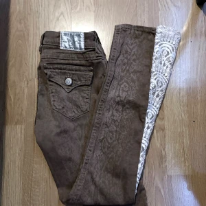 Bruna mönstrade jeans med spets - Säljer ett par bruna jeans från True Religion med diskret mönster och unik vit spetsdetalj längs ena benet. Klassisk femficksmodell med dekorativa sömmar och silverfärgade knappar. Perfekta för dig som vill sticka ut lite extra! Storlek 27. Sytt bootcut