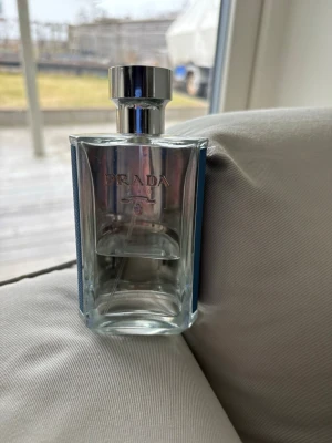 Prada L'Homme Eau de Toilette - Elegant parfym från Prada i en stilren, genomskinlig glasflaska med silverfärgat lock och blått textilband på sidan. Flaskans design är modern och exklusiv, perfekt för dig som gillar lyxiga dofter och snygga detaljer i badrumshyllan.  Ca 50% kvar.