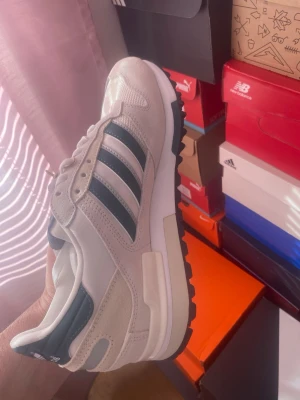 Adidas Originals vita sneakers - Fräscha Adidas Originals sneakers i vitt med svarta och mörkgröna detaljer samt klassiska tre ränder på sidan. Ovandelen är i en mix av mocka och syntetmaterial, med vit sula och svart yttersula. Perfekta för en sportig och stilren look.