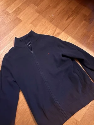 Svart ziptröja från Tommy Hilfiger - Snygg svart ziptröja från Tommy Hilfiger i ekologisk bomull. Tröjan har hög krage, hel dragkedja och en liten broderad logga på bröstet. Perfekt för en stilren och avslappnad look.