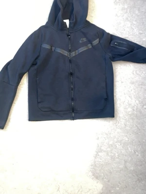 Mörkblå Nike hoodie med dragkedja - Snygg mörkblå hoodie från Nike med hel dragkedja och huva. Har två stora fickor framtill och en extra ficka med dragkedja på ena ärmen. Sportig design med diskreta svarta detaljer över bröstet.
