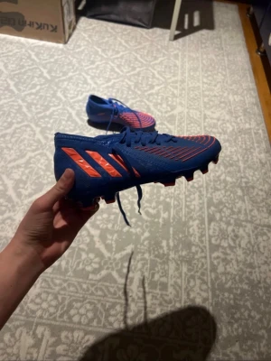 Blå Adidas Predator fotbollsskor - Säljer ett par blå Adidas Predator Edge fotbollsskor med röda detaljer och klassiska tre ränder på sidan. Skorna har snörning och är tillverkade i syntetmaterial för bra grepp och kontroll på planen. Perfekta för dig som vill sticka ut på matchen!