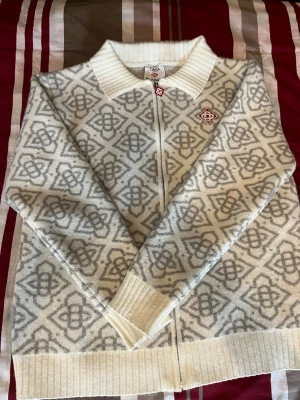 Vit och grå mönstrad kofta från CASA BLANCA - Snygg vit och ljusgrå kofta från CASA BLANCA med klassiskt geometriskt mönster. Dragkedja framtill, ribbstickad krage, muddar och nederkant. Broderad logotyp på bröstet. Perfekt för dig som gillar skandinavisk design.