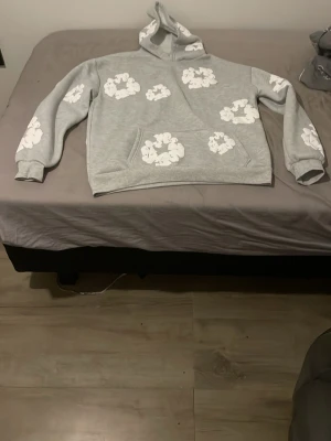 Grå hoodie från Denim Tears - Snygg grå hoodie från Denim Tears med vita blomliknande tryck över hela tröjan. Klassisk känguruficka framtill och huva. Perfekt för dig som vill sticka ut med en unik och trendig stil. Helt ny säljer den för den är för stor för mig 