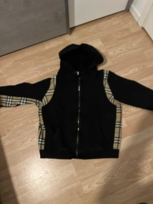 Burberry hoodie  - Svart hoodie med dragkedja och huva. Snygga beige-rutiga paneler längs ärmar och sidor som ger en klassisk streetstyle-vibe. Perfekt för dig som gillar en mix av basic och mönstrat. Mjuk bomullskänsla och två fickor framtill.