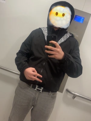 Svart vindjacka - Svart vindjacka med dragkedja och huva. Insidan av huvan och ärmarna har ett vitt och svart mönster med bokstäver. Jackan har elastiska muddar och en sportig look, perfekt för streetwear. Materialet är tunt och vindtätt