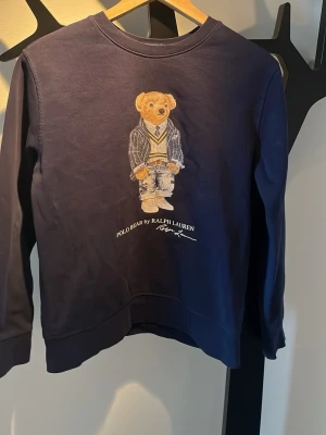 Marinblå hoodie Polo Ralph Lauren - Säljer en marinblå hoodie från Polo Ralph Lauren med den ikoniska Polo Bear-björnen broderad på bröstet. Hoodien har långa ärmar och rund halsringning. Perfekt för dig som vill ha en klassisk och stilren look med en twist.