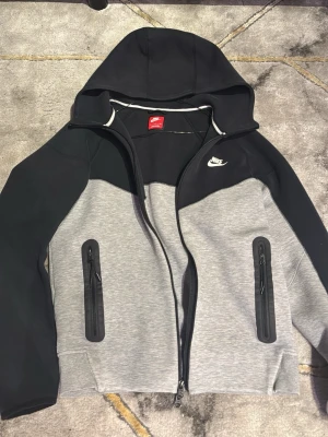Nike grå och svart hoodie med dragkedja - Nike hoodie i grått och svart med hel dragkedja framtill, två fickor med dragkedja och vit Nike-logga på bröstet. Hoodien har huva och är tillverkad i mjukt material, perfekt för en sportig och avslappnad stil.