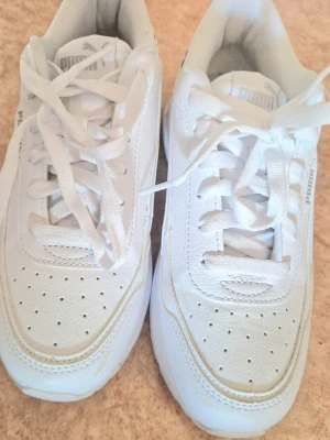 Vita sneakers från Puma - Säljer ett par helvita sneakers från Puma med klassisk design. Skorna har lågt skaft, perforerade detaljer på tån och sidorna samt vita skosnören. 