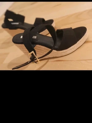 Svarta kilklackade espadrillesandaler - Snygga svarta sandaler med kilklack och flätad sula i espadrillestil. Remmar i mockaimitation och justerbart spänne i guld. Perfekta för sommarens alla outfits och ger extra höjd med bekväm känsla.