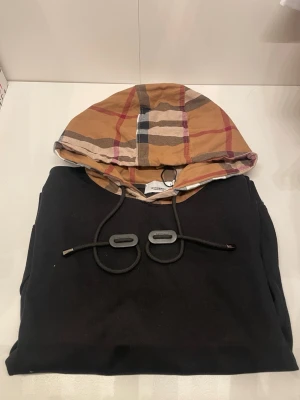 Svart hoodie med mönstrad huva - Svart hoodie med klassisk känguruficka och dragsko i halsen. Huvan har ett snyggt rutigt mönster i beige, rött och blått som ger en unik touch. Perfekt för dig som vill ha en stilren men ändå speciell hoodie till vardags.