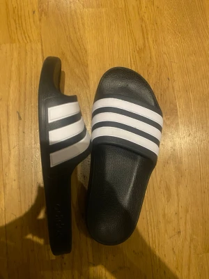 Svarta Adidas badtofflor med vita ränder - Säljer ett par klassiska svarta Adidas badtofflor med tre vita ränder över foten. Tillverkade i syntetmaterial med en bekväm och formad sula. Perfekta för stranden eller gymmet och har den ikoniska sportiga looken.