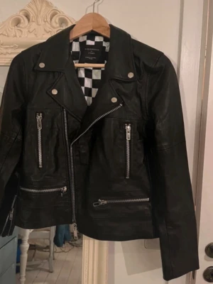 Svart skinnjacka Deadwood x Lindex - Snygg svart skinnjacka från Deadwood for Linc&Co i klassisk bikerstil med silvriga dragkedjor och tryckknappar. Jackan har schackrutigt foder och flera fickor med dragkedja. Perfekt för dig som vill ha en edgy look. Ett måste i varje fashionistas basgarderob! Stlk 36/38 men den är liten i storleken. Aldrig använd då den är för liten. Nypris ca 4000:-!