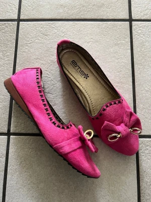 Rosa ballerinaskor med rosett ENS - Snygga ballerinaskor i starkt rosa mockaimitation från ENS Fashion. Skorna har dekorativ rosett framtill med guldfärgade detaljer och svarta sömmar längs kanten. Perfekta för dig som vill sticka ut med färg och stil.