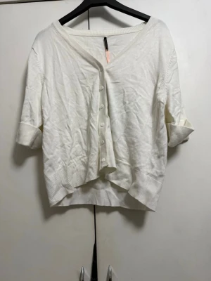 Vit kofta med knappar och v-ringning - Snygg vit kofta med v-ringning och knäppning framtill. Modellen har halvlånga ärmar med uppvik och dekorativa knappar vid ärmslutet. Ribbstickade muddar vid ärmar och nederkant. Perfekt att slänga över en topp för en chill look.