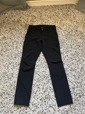  Trekmates byxor S - Säljer ett par svarta cargobyxor från Trekmates med raka ben och dragkedjeförsedda fickor på sidorna. Byxorna har bälteshällor och knappstängning i midjan. Tillverkade i ett slitstarkt och flexibelt material som passar perfekt för äventyr och vardag.