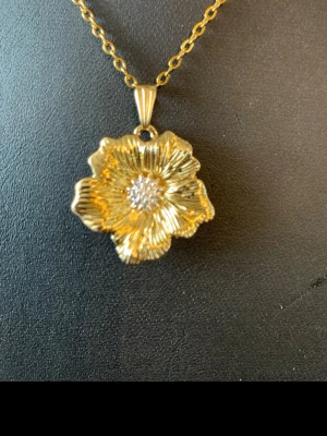 Guldigt halsband med blomma - Snyggt halsband med en guldfärgad blomformad berlock och små glittrande stenar i mitten. Kedjan är tunn och elegant, perfekt för att ge din outfit en extra touch. Blomman har fina detaljer och en lyxig känsla.