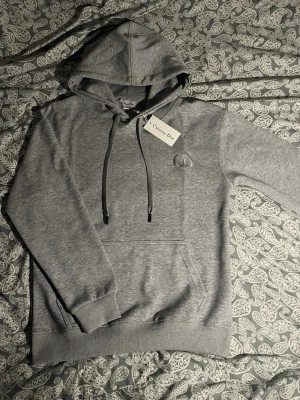 Grå hoodie - Snygg grå hoodie från med broderad CD-logga på bröstet. Hoodien har huva med dragsko, ribbade muddar och magficka. Tillverkad i mjukt bomullsmaterial som känns skönt mot huden. Perfekt för en avslappnad och trendig look.