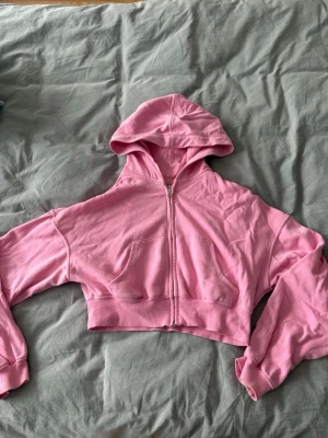 Rosa cropped hoodie med dragkedja - Säljer en rosa cropped hoodie med dragkedja framtill och huva. Hoodien har två fickor på magen och breda muddar vid ärmslut och midja. Perfekt för en chill och avslappnad stil.