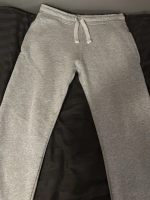 Grå mjukisbyxor russemerch - Snygga grå mjukisbyxor med dragsko i midjan och fickor på sidorna samt en bakficka. Byxorna har en klassisk rak passform och är gjorda i mjukt och bekvämt material, perfekt för chill dagar. Enkla att matcha med hoodie eller t-shirt.