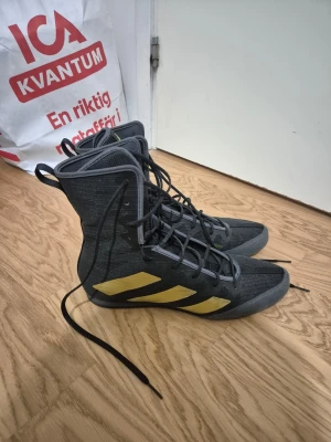 Adidas svarta boxningsskor med guld - Helt oanvända skor! De är i storlek 44 men passade mig bra och jag har storlek 43 eftersom de är små i storleken.                                                                                                    Säljer ett par svarta höga boxningsskor från Adidas med guldiga ränder på sidorna och guldigt Adidas-logga på plösen. Skorna har snörning hela vägen upp och är tillverkade i ett lätt och ventilerande material som passar perfekt för träning och tävling.