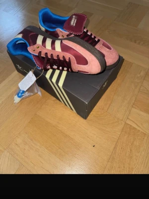 Adidas burgundy och rosa sneakers - Snygga Adidas sneakers i vinrött och rosa mocka med klassiska vita ränder på sidorna. Skorna har svart sula, blå insida och detaljer i gult vid snörningen. Perfekt för dig som vill sticka ut med färg och retrostil. Levereras med originalkartong.