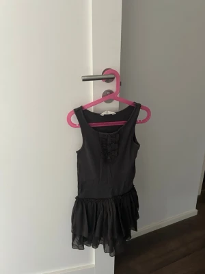  kort klänning från H&M - Säljer en svart kort klänning från H&M med ärmlös design och volangdetaljer på kjolen. Framtill finns dekorativa rynkningar som ger en söt touch. Perfekt för dig som gillar enkel men stilren look.
