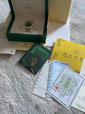 Elegant Role Klocka i Stål med Grön Urtavla – Komplett Set - Säljer en mycket stilren Role klocka med exklusiv grön urtavla och klassiskt tvåtonsarmband i guld och stål. Den räfflade guldringen tillsammans med den djupgröna tavlan ger klockan ett lyxigt och tidlöst utseende som passar både till vardag och fest.  Klockan säljs som komplett set och kommer med box, ytterbox, certifikat, manualer, tagg och tillbehör enligt bilder. Den är väl omhändertagen och förvarad i sin originalbox.  Ingår: • Innerbox och ytterbox • Certifikat / garantikort • Manualer och