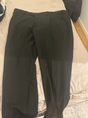 Svarta kostymbyxor med raka ben - Svarta kostymbyxor med klassisk rak passform och hällor för skärp. Byxorna har knäppning i midjan och fickor både fram och bak. Perfekta för en stilren look och passar till skjorta eller t-shirt.