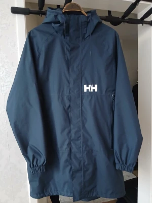 Mörkblå regnjacka från Helly Hansen - Snygg mörkblå regnjacka från Helly Hansen med huva och dragkedja framtill. Jackan har elastiska muddar vid ärmsluten och Helly Hansen-logga på bröstet. Tillverkad i vattentätt material, perfekt för regniga dagar.