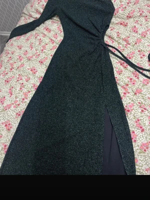 Glittrig mörk grön långklänning med slits - Snygg svart långklänning med glittrigt tyg och lång ärm. Klänningen har en markerad midja med knyt och en hög slits framtill som ger en extra touch. Perfekt för dig som vill sticka ut med stil.