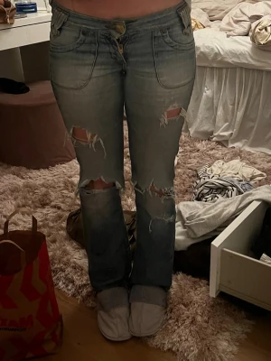 Ljusblå slitna bootcut jeans - Säljer ett par ljusblå bootcut jeans med flera slitningar och hål på benen. Jeansen har klassiska fem fickor, låg midja och snygga detaljer på bakfickorna. Perfekta för en avslappnad och trendig look. Jag är 169 cm och de passar perfekt💗