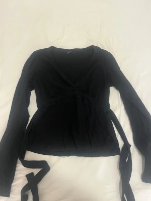 Svart omlottopp med långa ärmar - Svart omlottopp med långa ärmar och knytband i midjan. Toppen har v-ringning och är tillverkad i ett mjukt, lätt ribbat material. Perfekt för en stilren och trendig look.