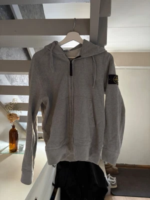 Grå zip hoodie från Stone Island - Snygg grå hoodie med dragkedja från Stone Island. Klassisk huva med dragsko och ikonisk logopatch på vänster ärm. Tillverkad i mjukt bomullsmaterial och har ribbade muddar vid ärmslut och nederkant. Perfekt för en avslappnad och trendig look.