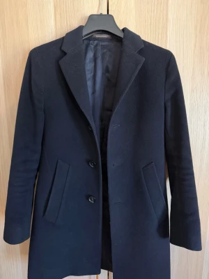 Oscar Jacobson Rock - Stilren svart kappa från Zara med rak passform, klassisk krage och knäppning framtill. Kappan har två snedställda fickor och är helfodrad för extra komfort. Perfekt för dig som gillar en tidlös look och vill hålla värmen under kyliga dagar.