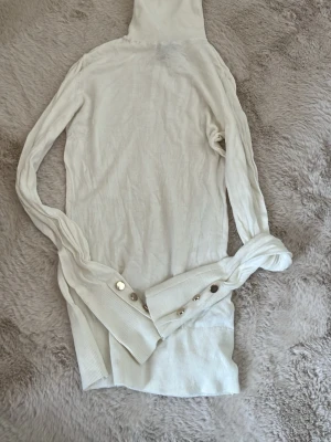 Vit stickad polotröja med knappar - Säljer en lång, vit stickad polotröja med snygga guldfärgade knappar på ärmarna. Tröjan har ribbade muddar och är tillverkad i mjukt stickat material. Perfekt för dig som gillar stilrena och trendiga plagg. sååå gullig!! notera att den är lite genomskinlig o påminner lite om intimissimi 😍 har en liknande i svart - pris är alltid diskuterbart❤️