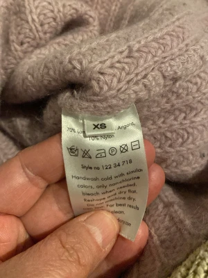 Ljuslila stickad tröja Hunkydory XS - Mysig ljuslila stickad tröja från Hunkydory i storlek XS. Tröjan är tillverkad av 70% lammull, 20% angora och 10% nylon, vilket gör den både mjuk och varm. Ribbstickad struktur och klassisk passform – perfekt för kyliga dagar.