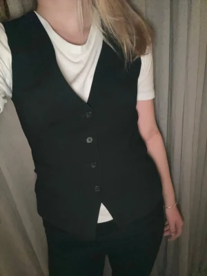 Svart klassisk väst med knappar - Stilren svart väst med v-ringning och fyra knappar framtill. Västen har en figurnära passform och är perfekt att styla över en t-shirt eller skjorta. Enkel och tidlös design som passar till många olika looks. 🩷🩷